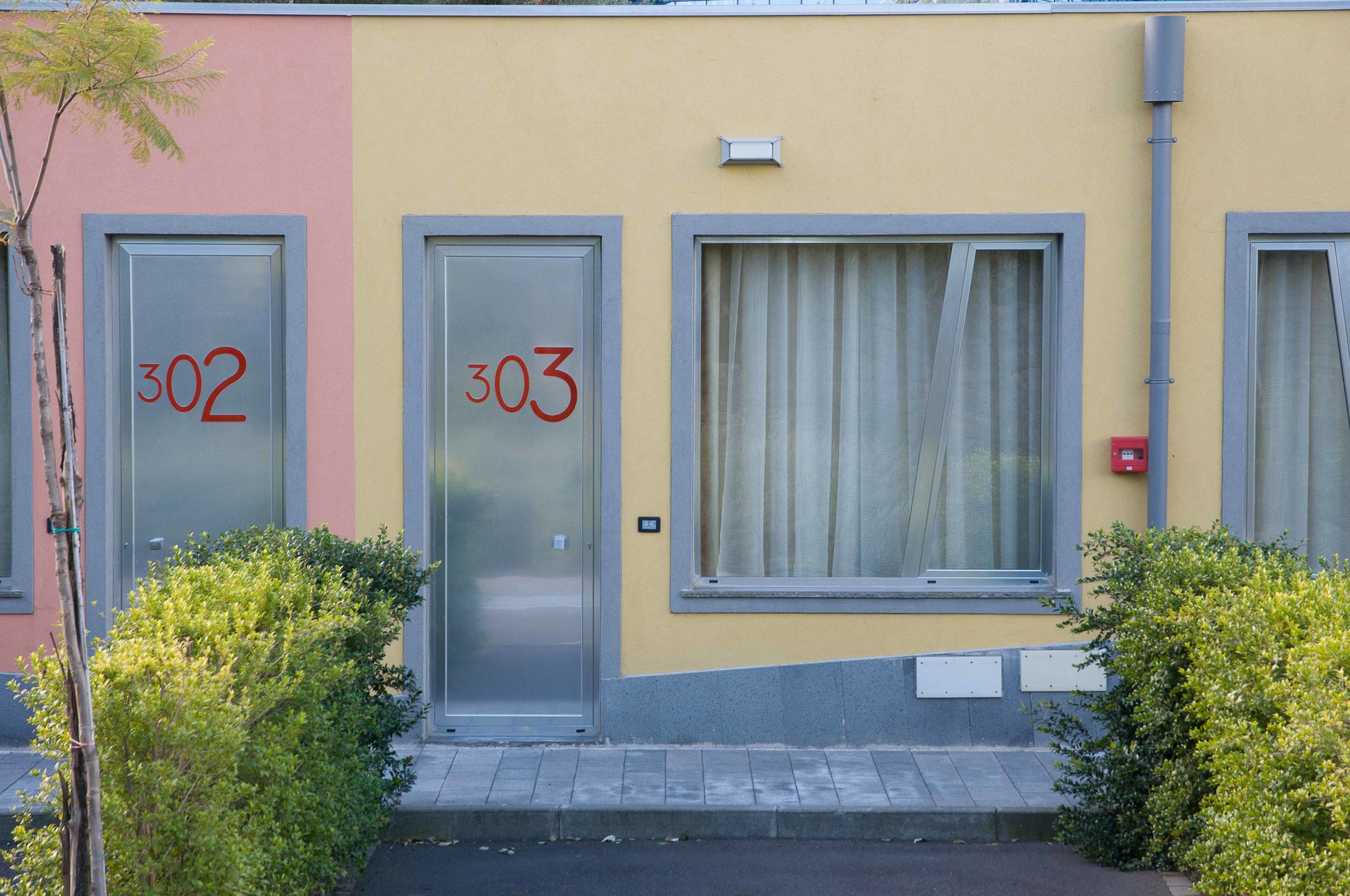 Un edificio con due porte, con le targhette 302 e 303, una finestra con tende e un piccolo spazio verde.