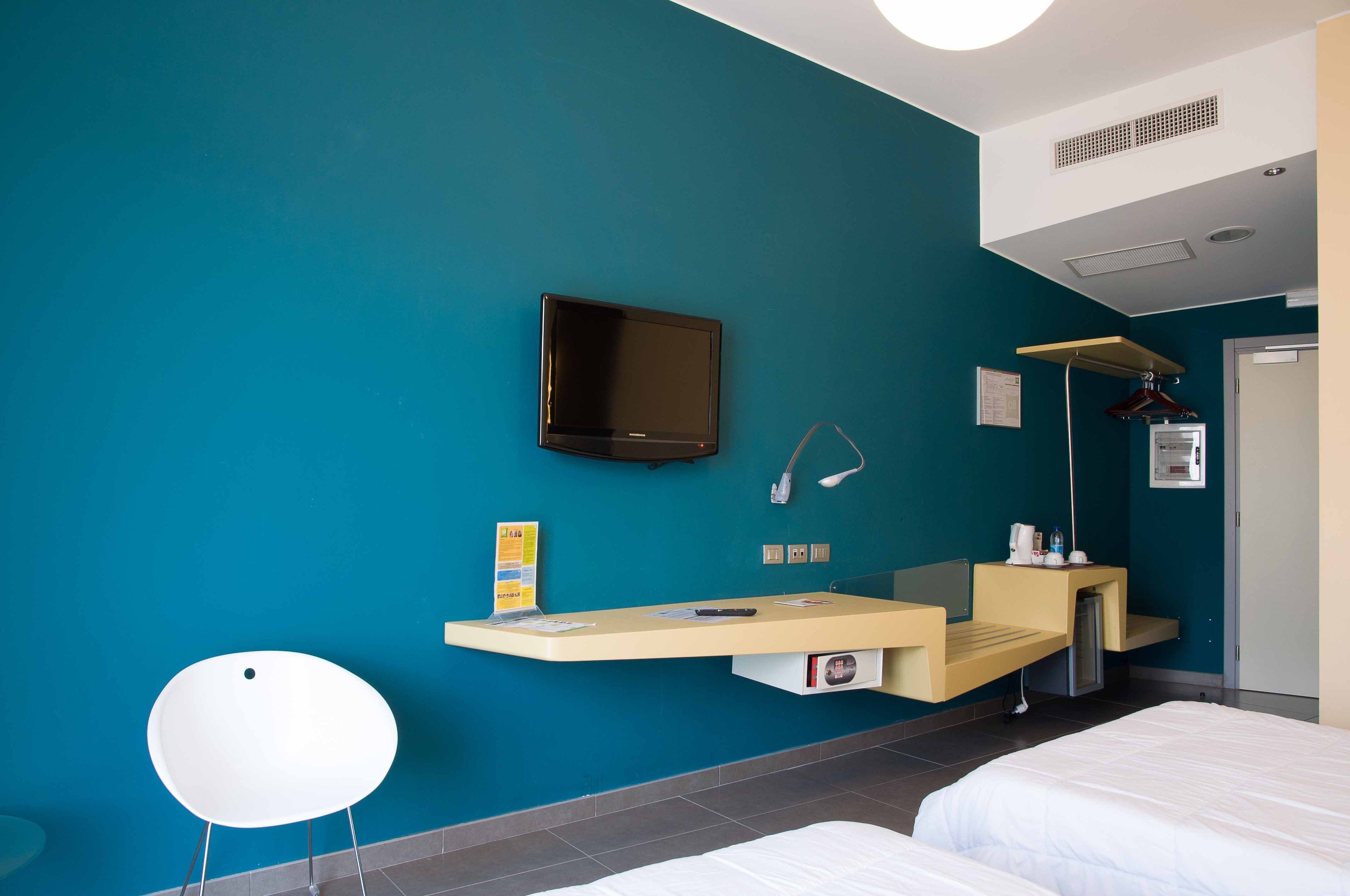 Ein Hotelzimmer mit einer türkisfarbenen Wand, einem flachbildfernseher über einem Schreibtisch, einem Bett und einem Kleiderschrank.