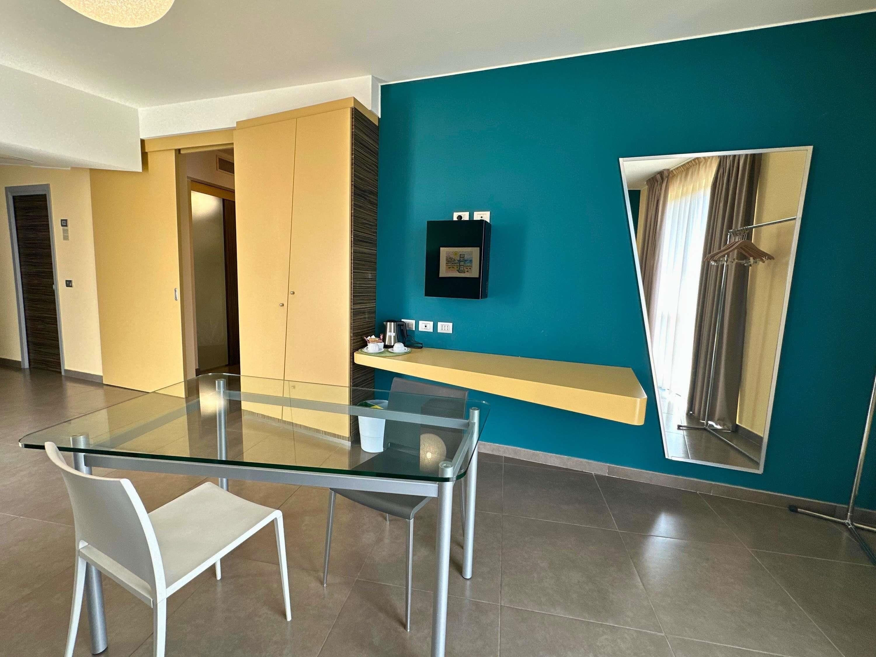 A modern hotel room with a glass dining table, a white chair, a built-in bench, a blue accent wall, and a large mirror.