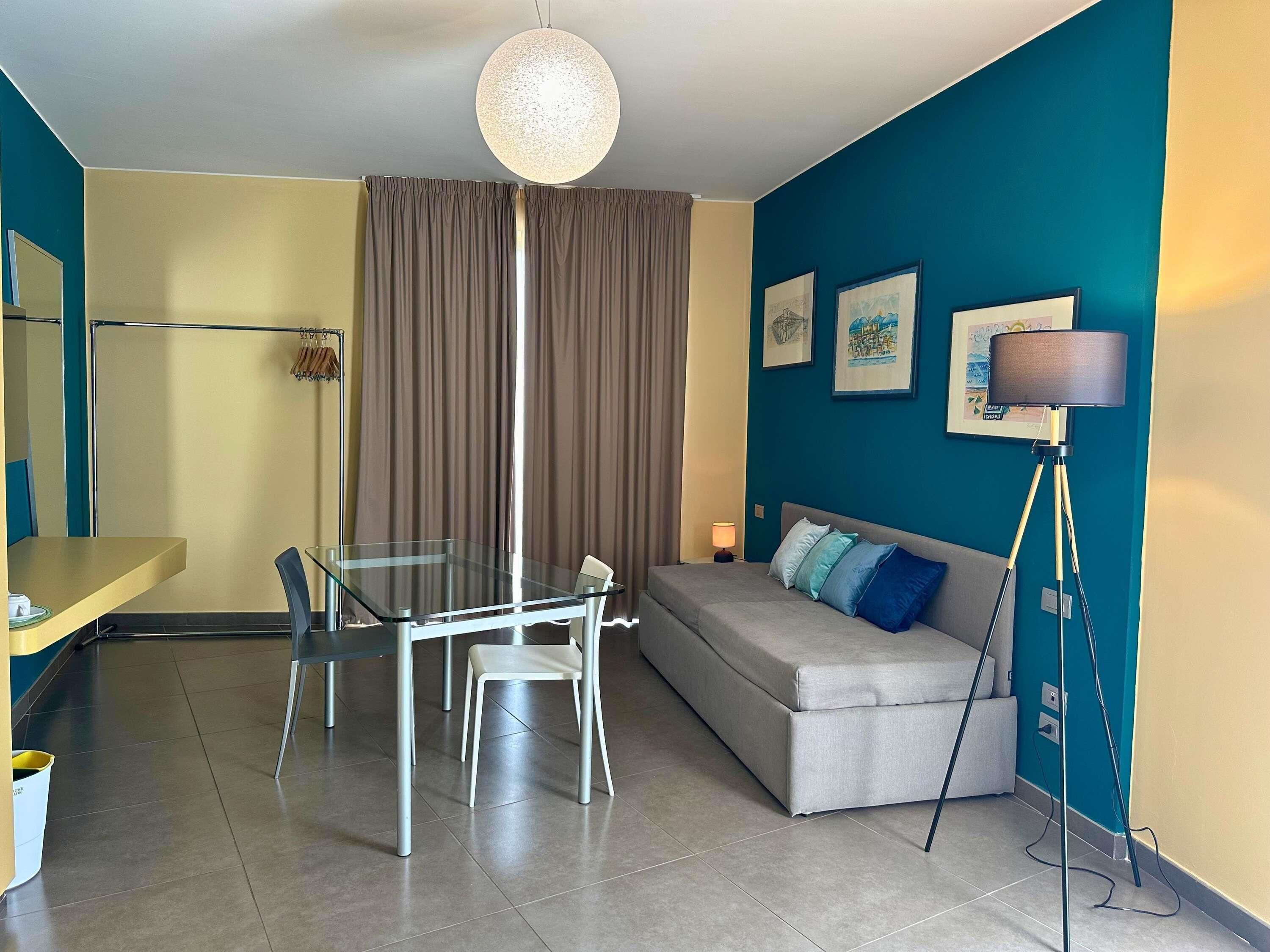 A modern living room with a glass dining table, a sofa with cushions, a standing lamp, and a ceiling light fixture.