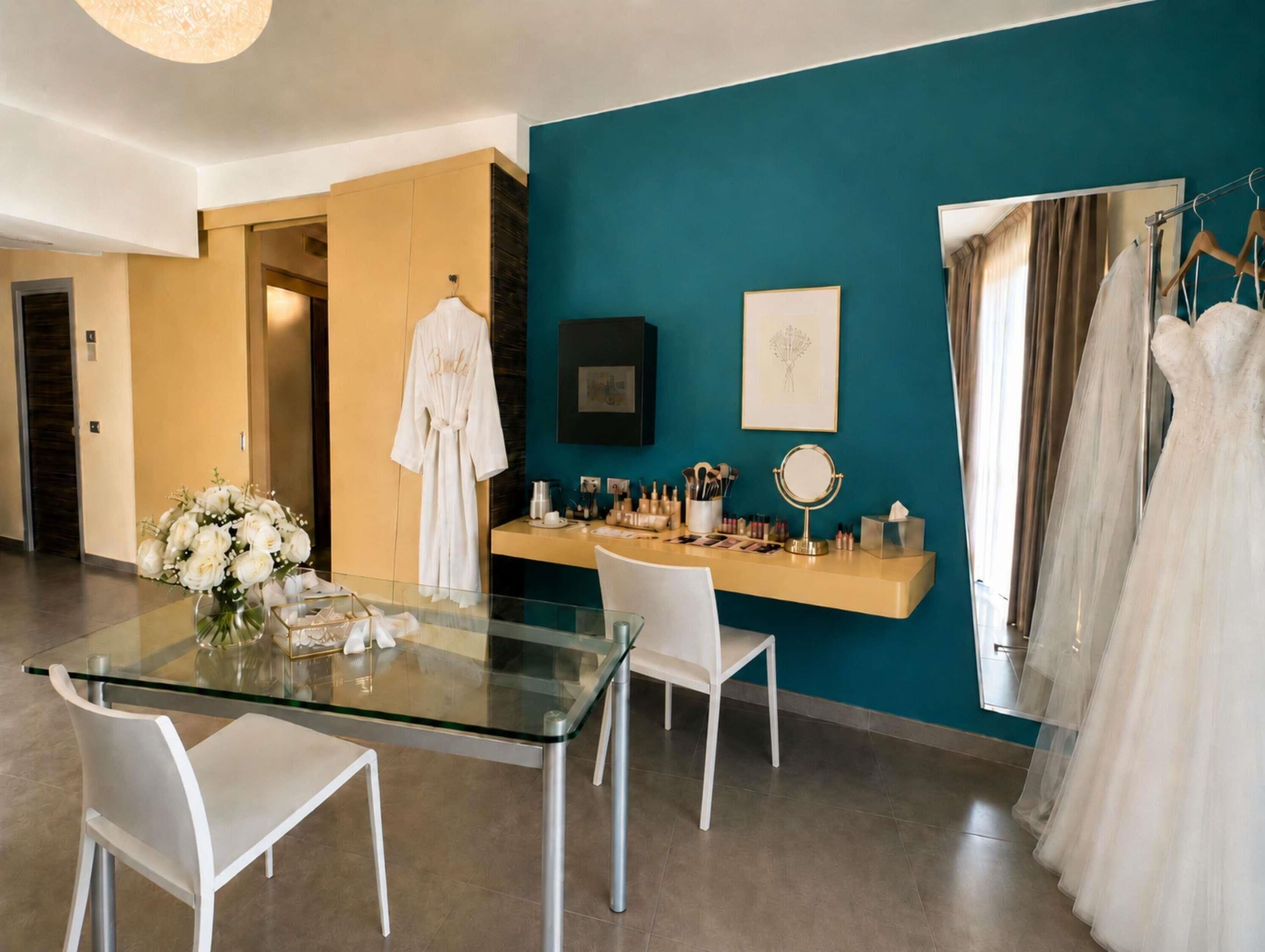 A modern hotel room with a glass dining table, white chairs, a vanity with makeup and accessories, and a wedding dress on a hanger.