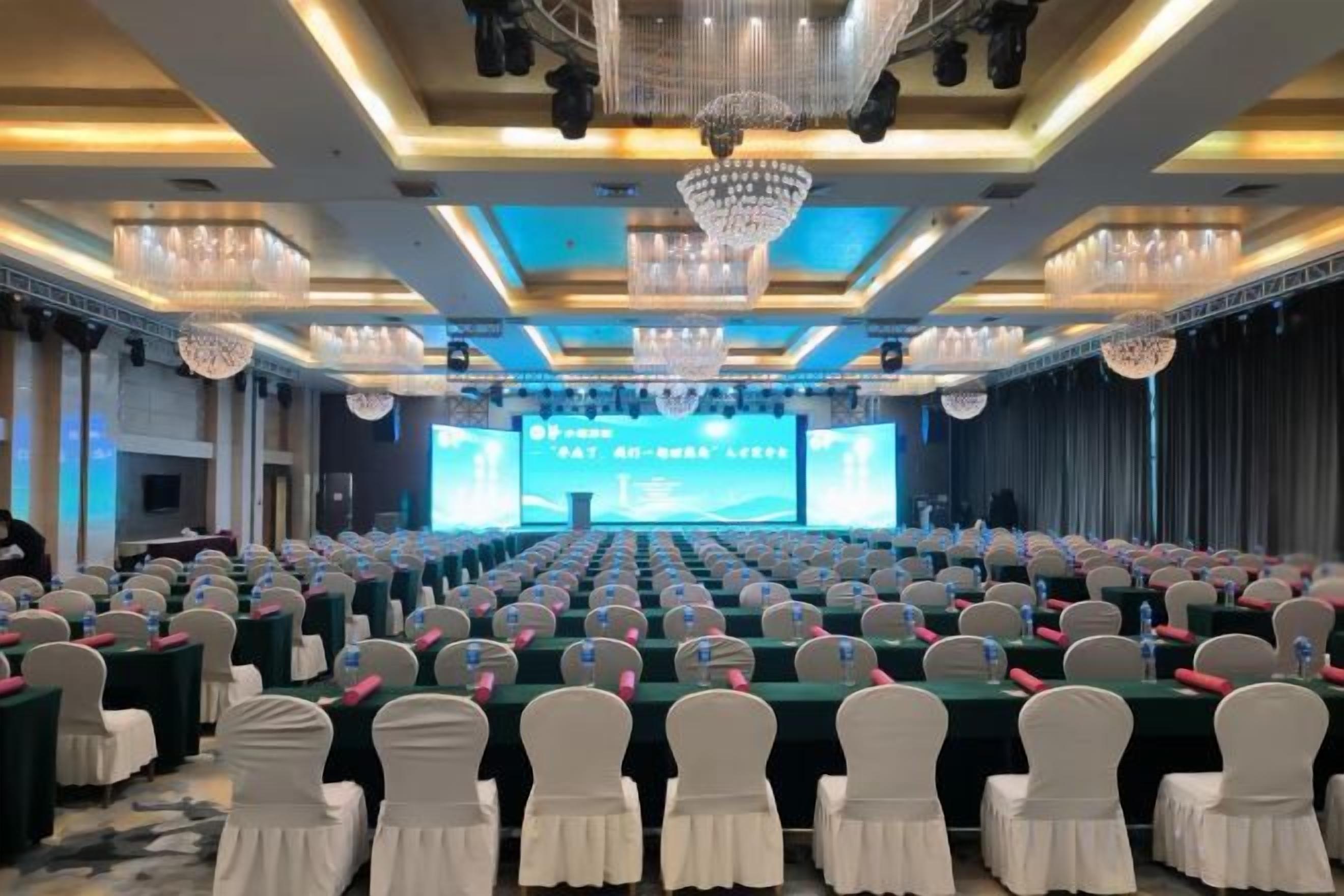A conference room with rows of tables, chairs, and a stage with a large screen.