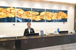 A hotel reception desk with a receptionist, a digital check-in kiosk, and a large floral artwork on the wall.