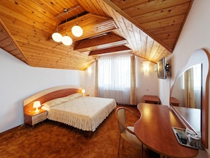 A bedroom with a wooden ceiling, a bed with a headboard, bedside lamps, a desk with a chair, a television, and a window with curtains. - Hotel Oasis (Brasov)