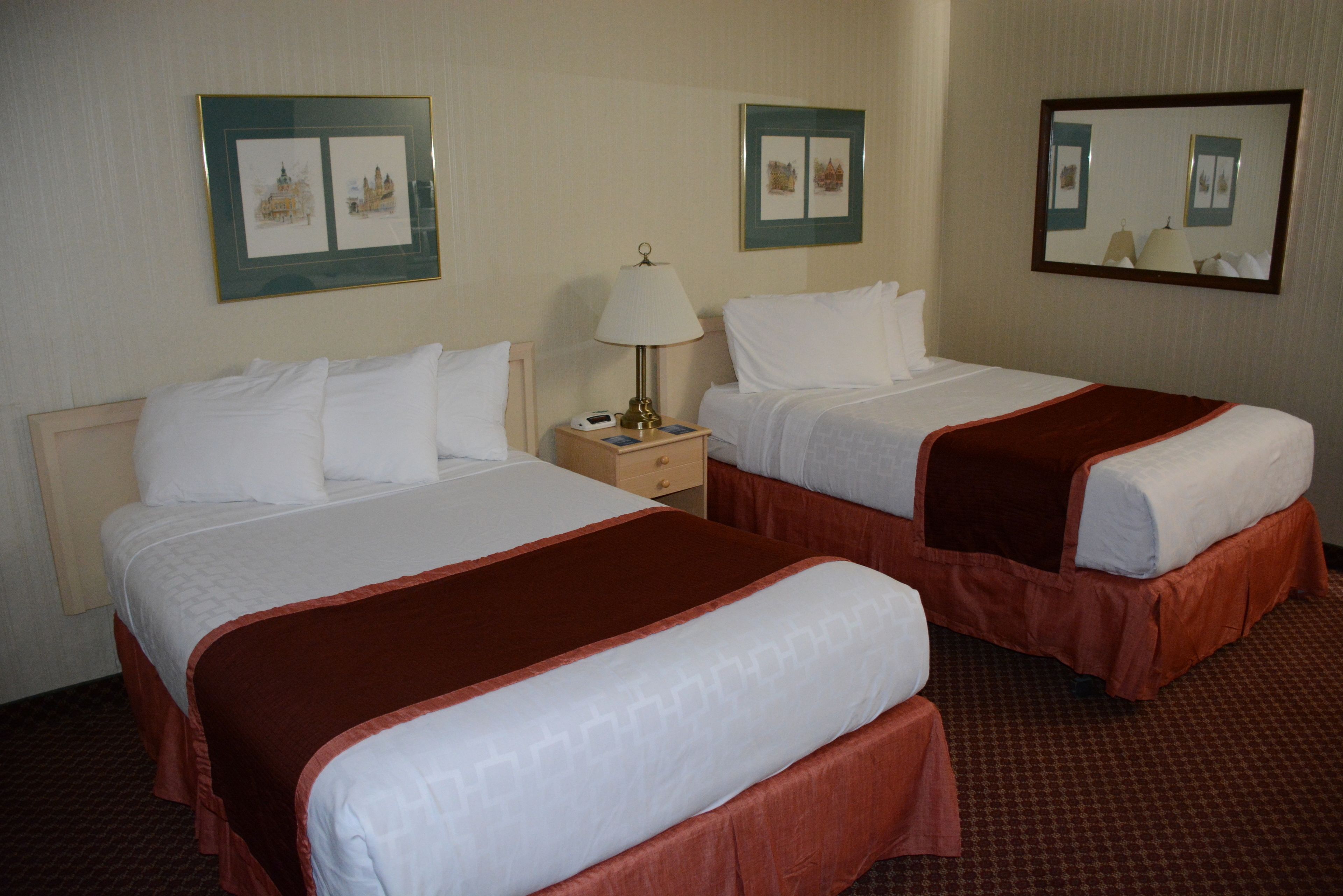 A hotel room with two beds, each with white bedding and a brown accent. There is a nightstand with a lamp between the beds.