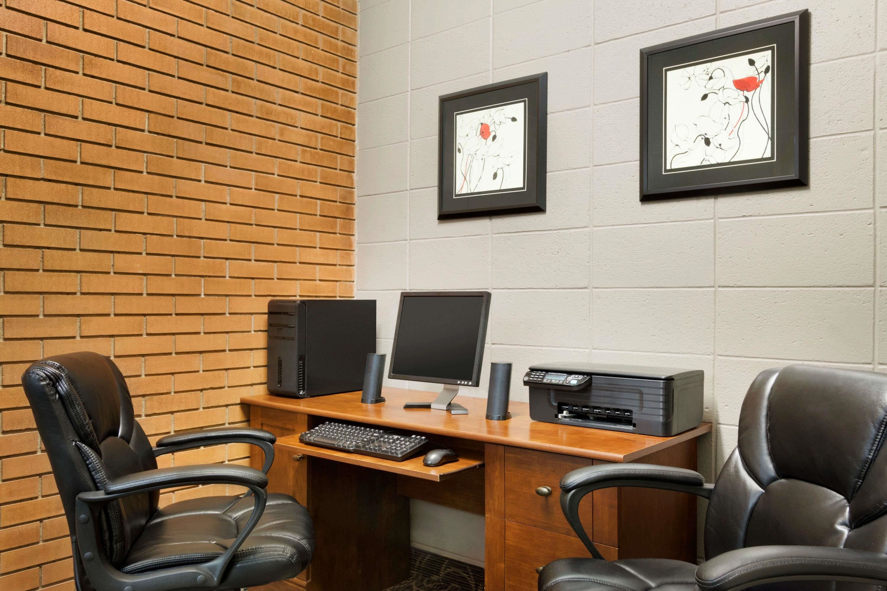 A conference room with a brick wall, two framed artworks, a desk with a computer, two chairs, and a printer.