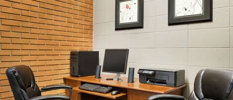 A conference room with a brick wall, two framed artworks, a desk with a computer, two chairs, and a printer.