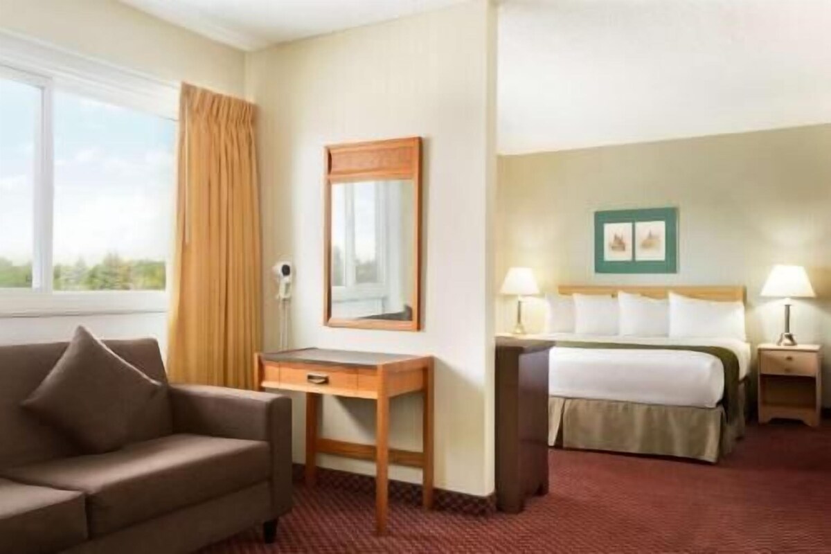A hotel room with a bed, a desk, a chair, a window with curtains, and a wall-mounted picture.