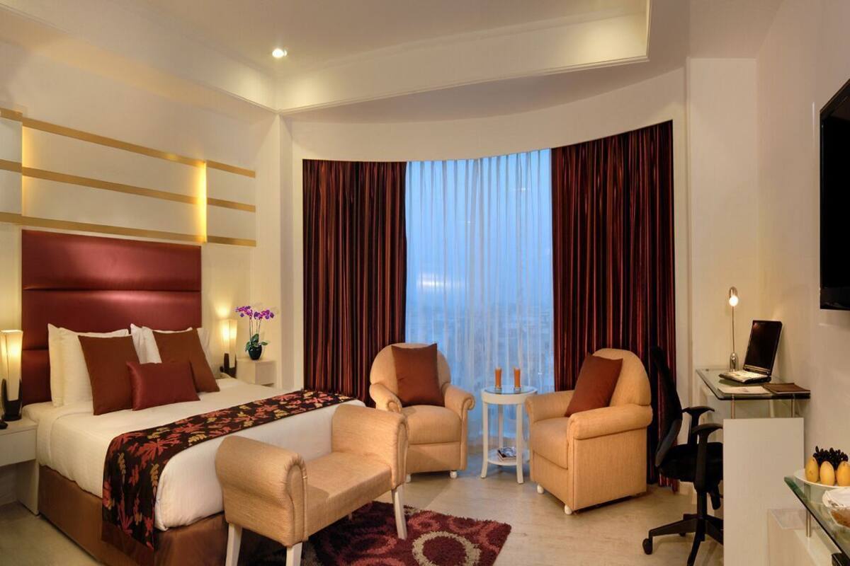 A hotel room with a bed, two armchairs, a small table, a desk with a laptop, and a TV.