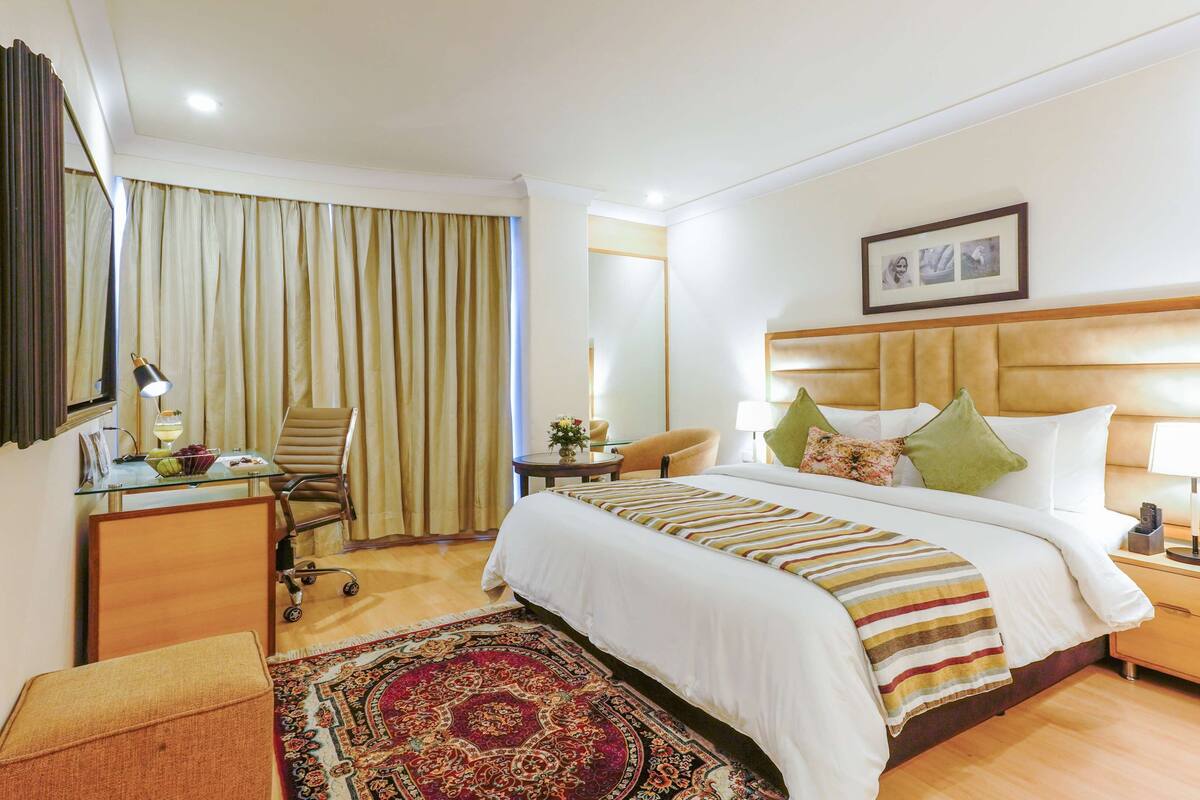A hotel room with a large bed, a desk with a lamp, a chair, a small table with a vase, and a patterned rug.