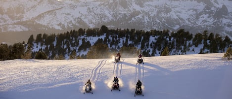 A group of four snowmobiles on a snowy mountain slope with a forested area and snow-covered peaks in the background.