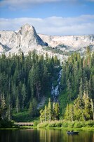Una baita con veranda che si affaccia su un lago, circondata da fitta foresta e con le montagne sullo sfondo.
