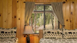 A wooden cabin bedroom with two beds, a nightstand, a lamp, and a window with curtains. - Tamarack Lodge and Resort (Mammoth Lakes)