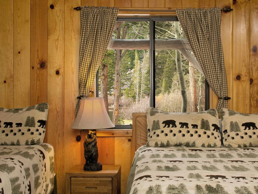 Un dormitorio tipo cabaña de madera con dos camas, una mesita de noche, una lámpara y una ventana con cortinas.