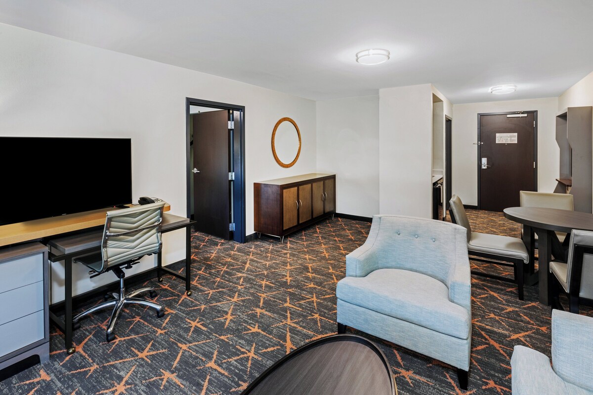A hotel room with a flat-screen TV, a desk with a chair, a sofa, a dining table with chairs, a wooden cabinet, and a patterned carpet.