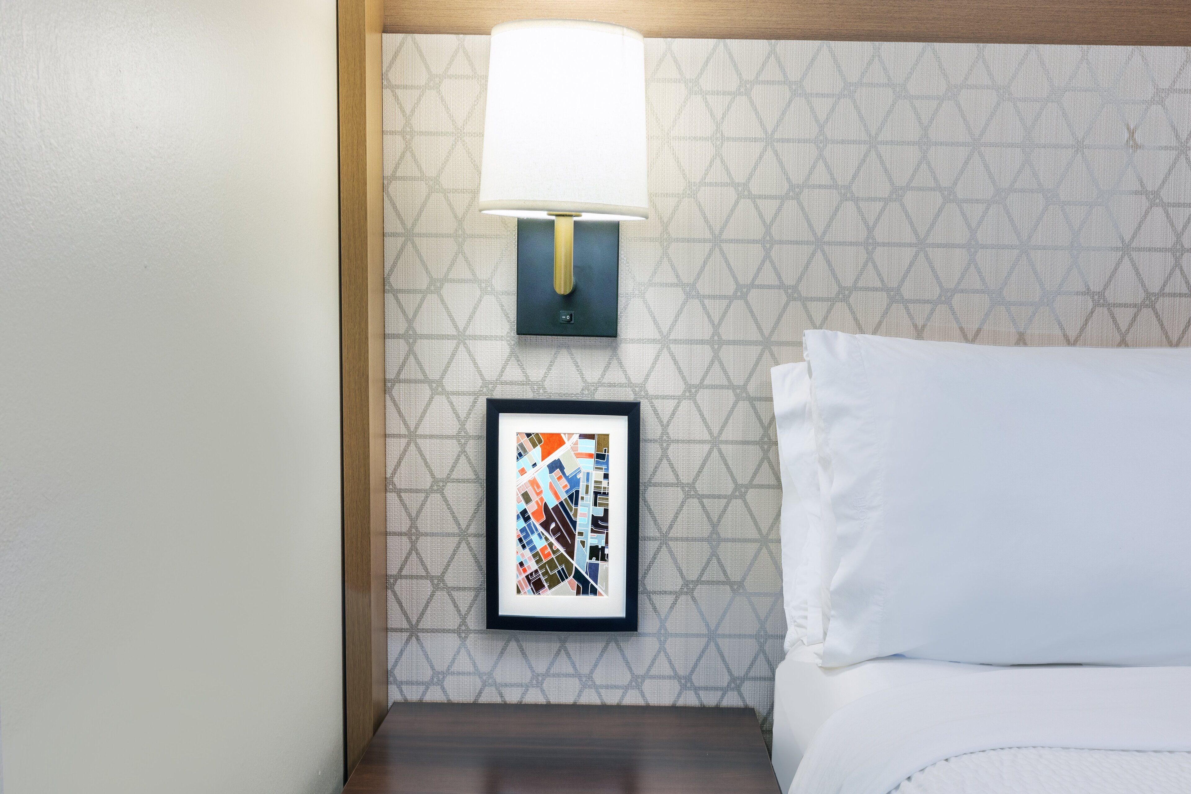 Cama bem arrumada com lençóis brancos, uma obra de arte abstrata emoldurada na parede e uma luminária fixada na parede.