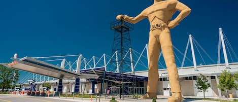 A large statue of a man with a hat and the word "TULSA" on his chest, standing in front of a modern building with a bridge in the background.
