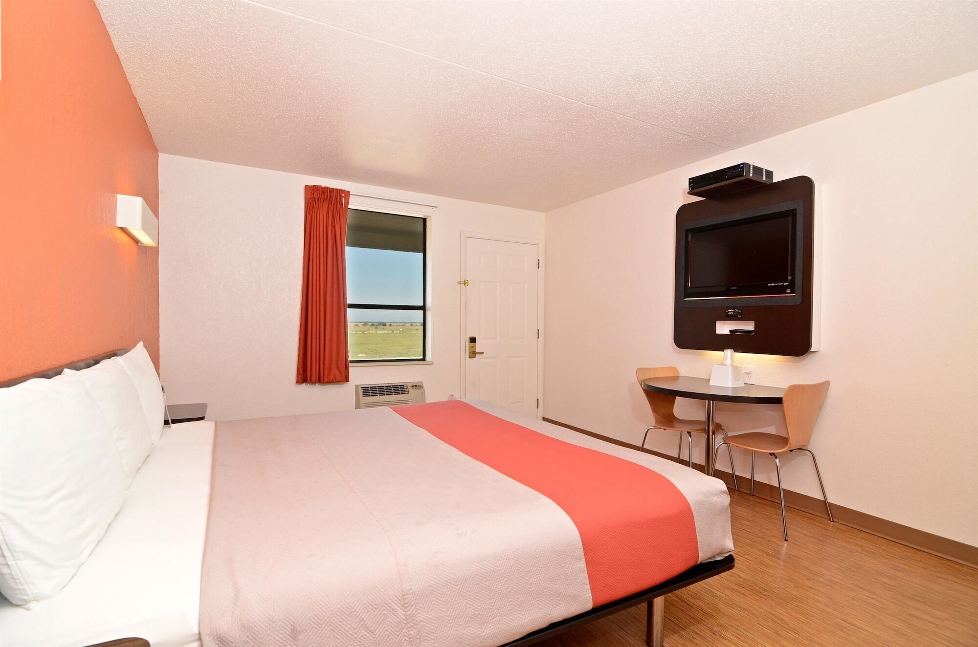 A hotel room with a bed, a desk with a chair, a television mounted on the wall, and a window with curtains.