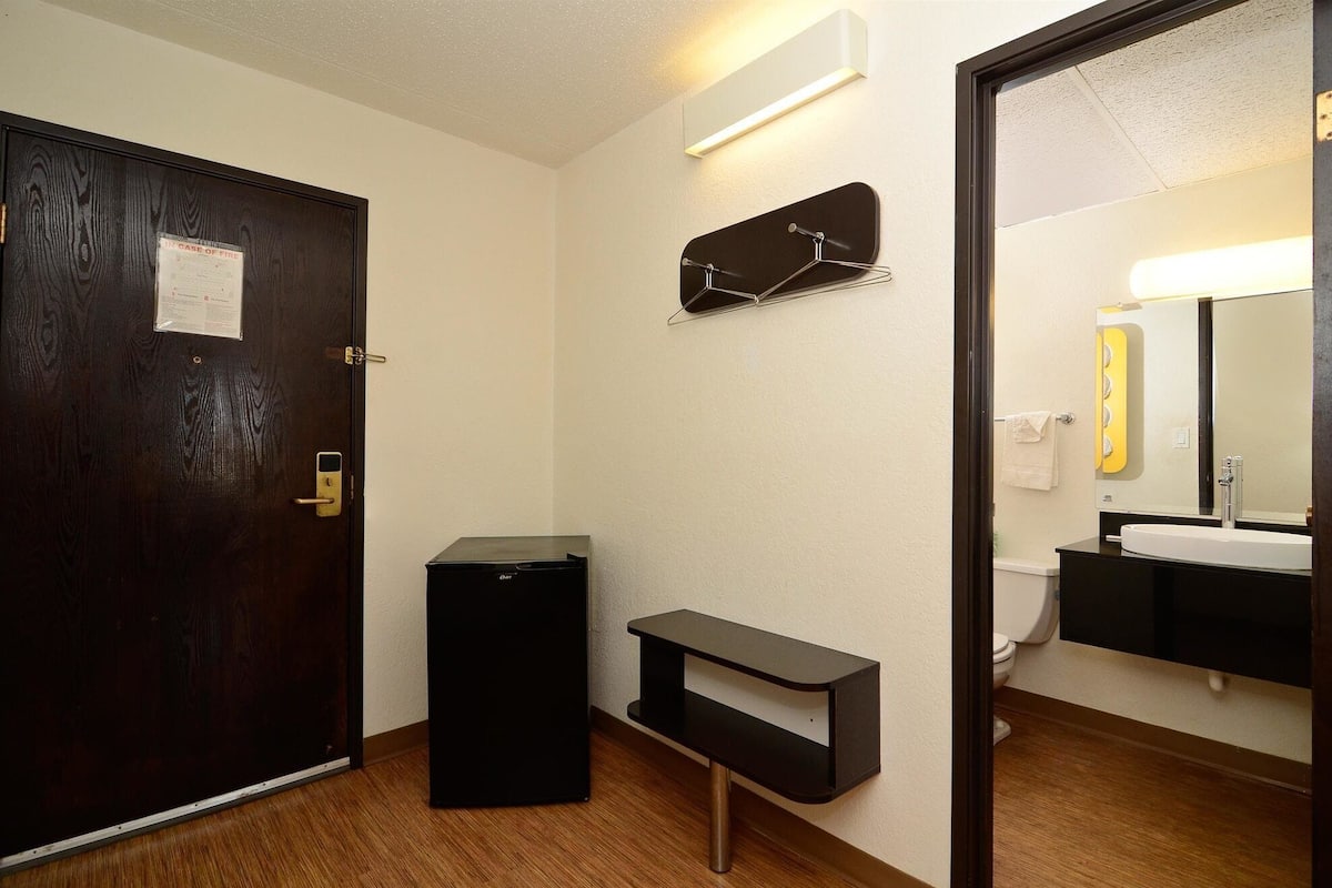 Un baño de hotel con una puerta de madera, un bote de basura negro, un estante pequeño negro y un espejo con marco amarillo.