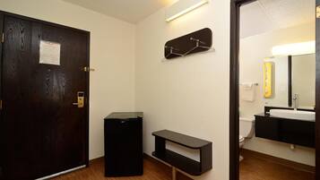 A hotel bathroom with a wooden door, a black trash bin, a small black shelf, and a mirror with a yellow frame.