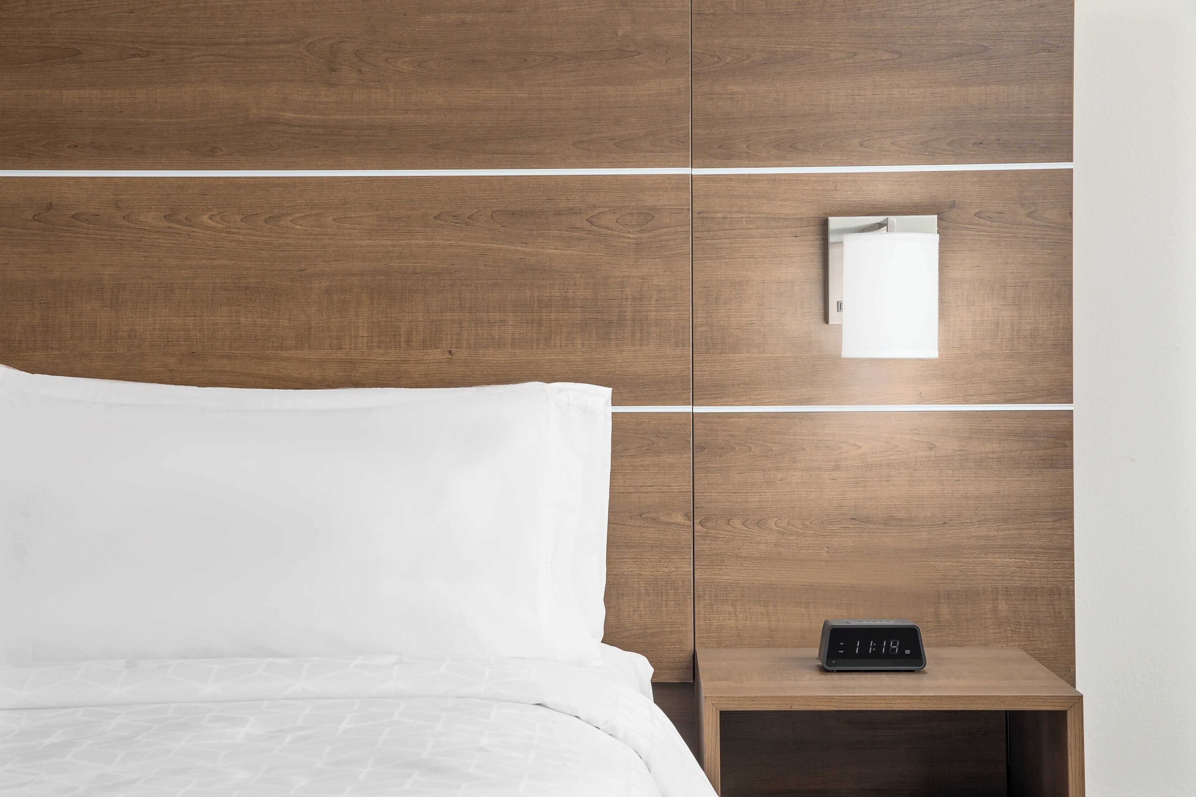 A neatly made bed with white linens, a wooden headboard, and a bedside table with a digital clock.