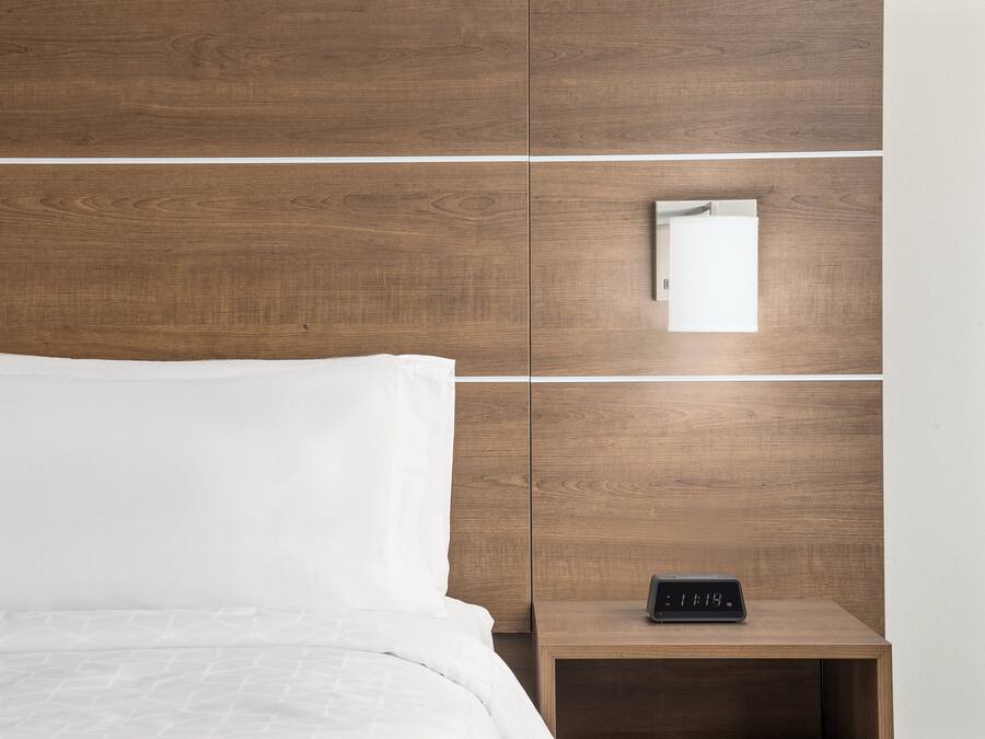 A neatly made bed with white linens, a wooden nightstand with a digital clock, and a mirror on the wall.