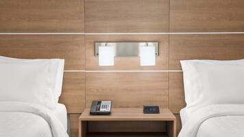 Habitación de hotel con cabecera de madera, dos camas con sábanas blancas, una mesita de noche con teléfono y un reloj digital, y un espejo sobre la mesita.