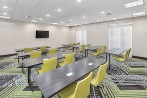 A conference room with multiple tables and chairs, a flat-screen TV on the wall, and patterned carpet.
