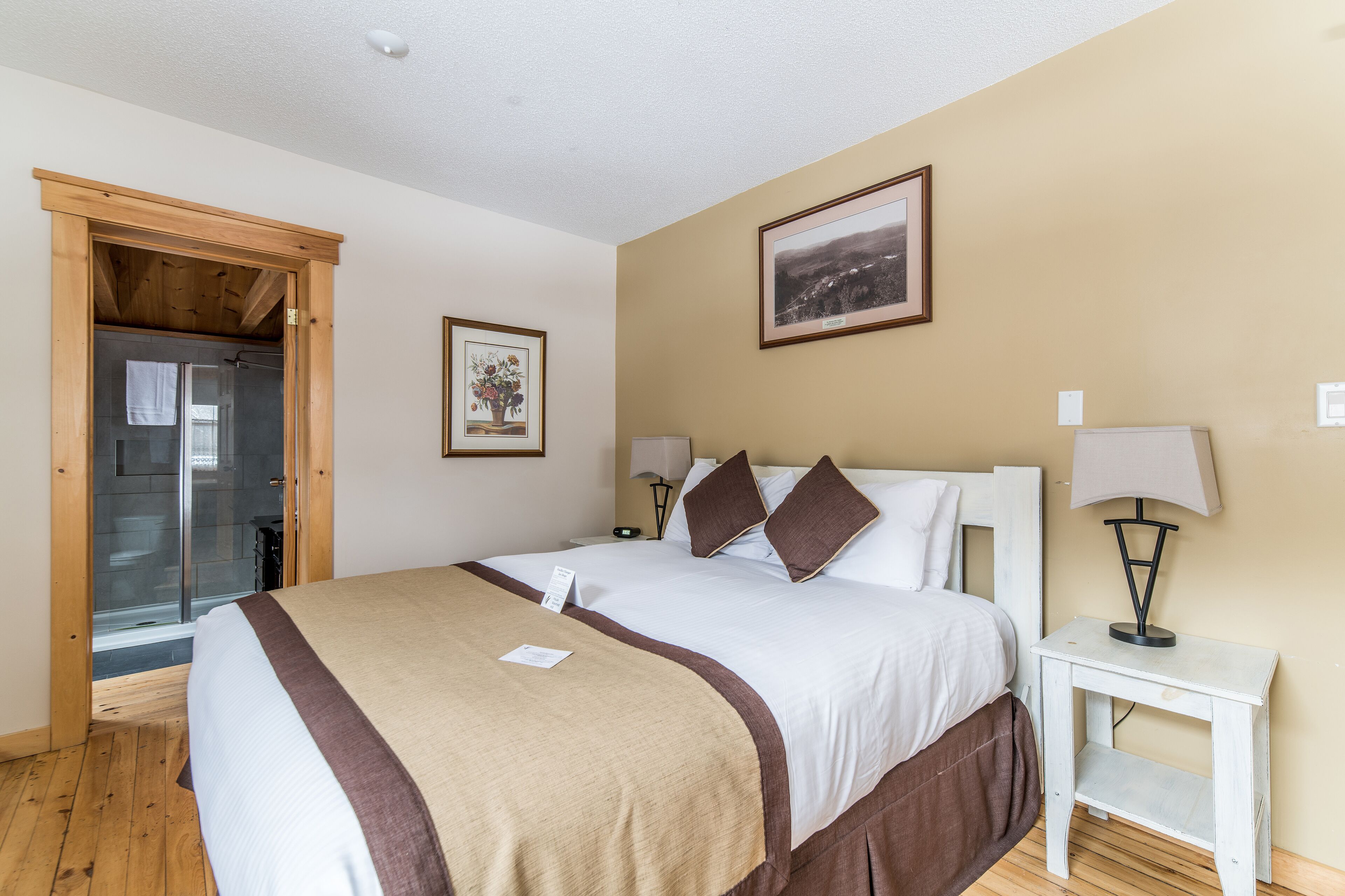 Una habitación de hotel con una cama grande, una puerta de madera que da a la zona de ducha, un cuadro en la pared y una pequeña mesa con una lámpara.