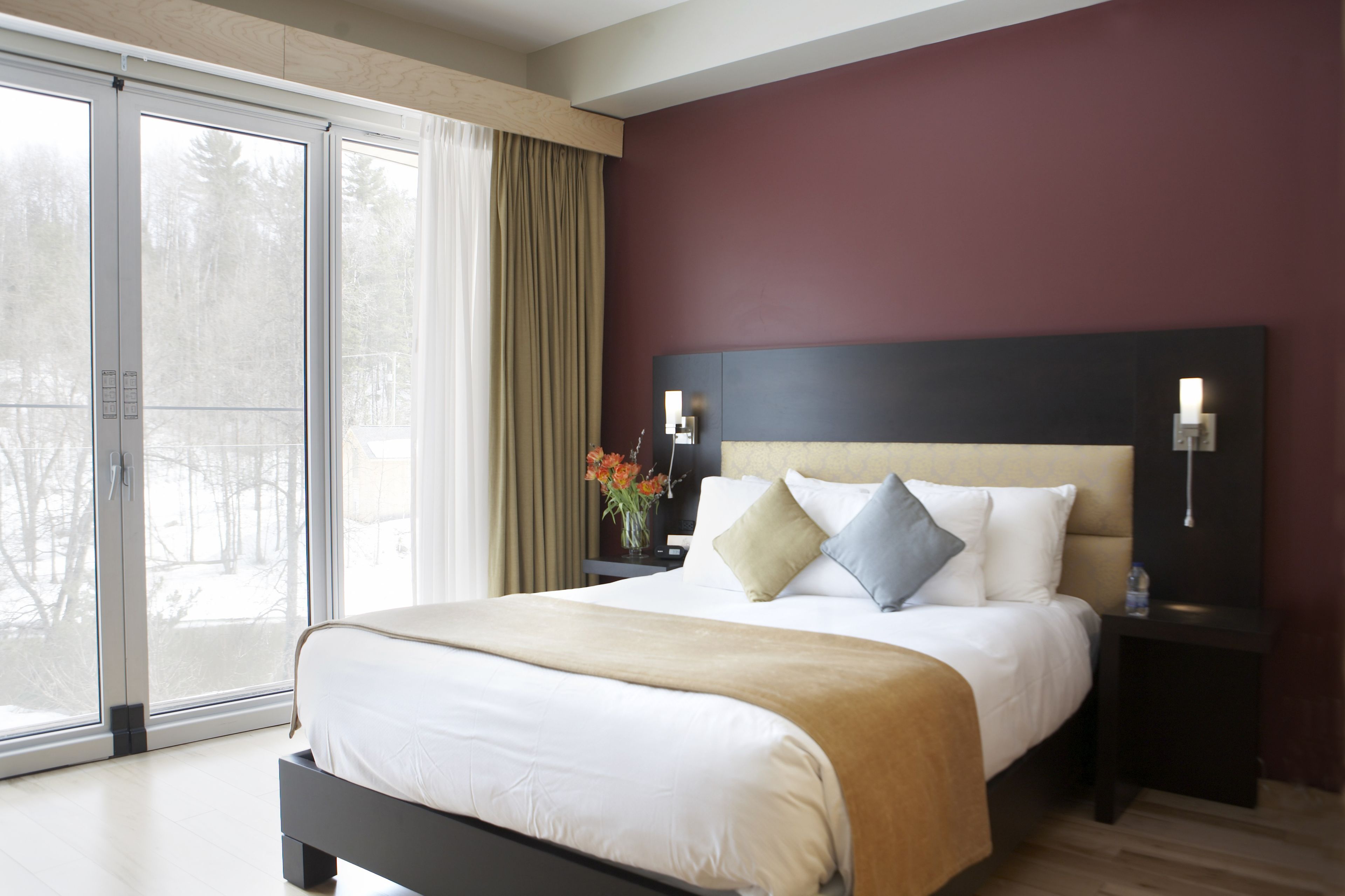 A modern hotel room with a large bed, a nightstand with a lamp, and a view through a sliding glass door.