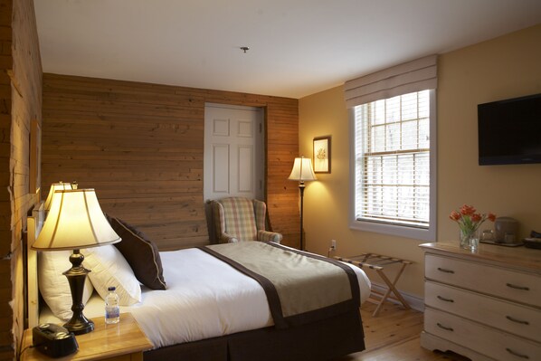 A hotel room with a wooden wall, a bed with a white and brown bedspread, a nightstand with a lamp, a chair, a dresser, a window with blinds, and a flat-screen TV mounted on the wall. - Moulin Wakefield Mill Hotel & Spa (La Peche)