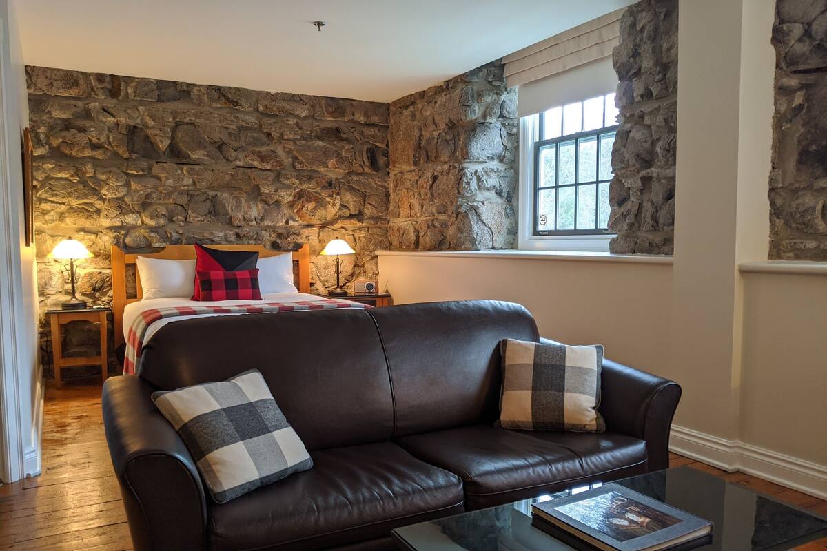Una habitación acogedora con una pared de piedra, una cama con un plaid rojo y negro, un sofá de cuero marrón con cojines cuadriculados y una mesa de centro de vidrio con un libro.