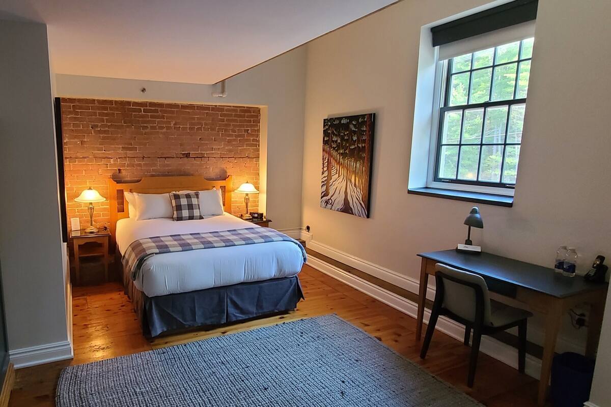 Un dormitorio con cabecera de ladrillo, cama con colcha escocesa, escritorio con silla, ventana con vistas a árboles y lámpara en la mesita de noche.