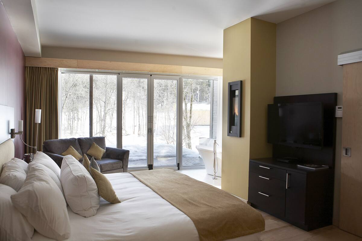 Habitación de hotel moderna con una cama grande, un televisor de pantalla plana, un sofá y vista a un paisaje nevado a través de puertas de vidrio.