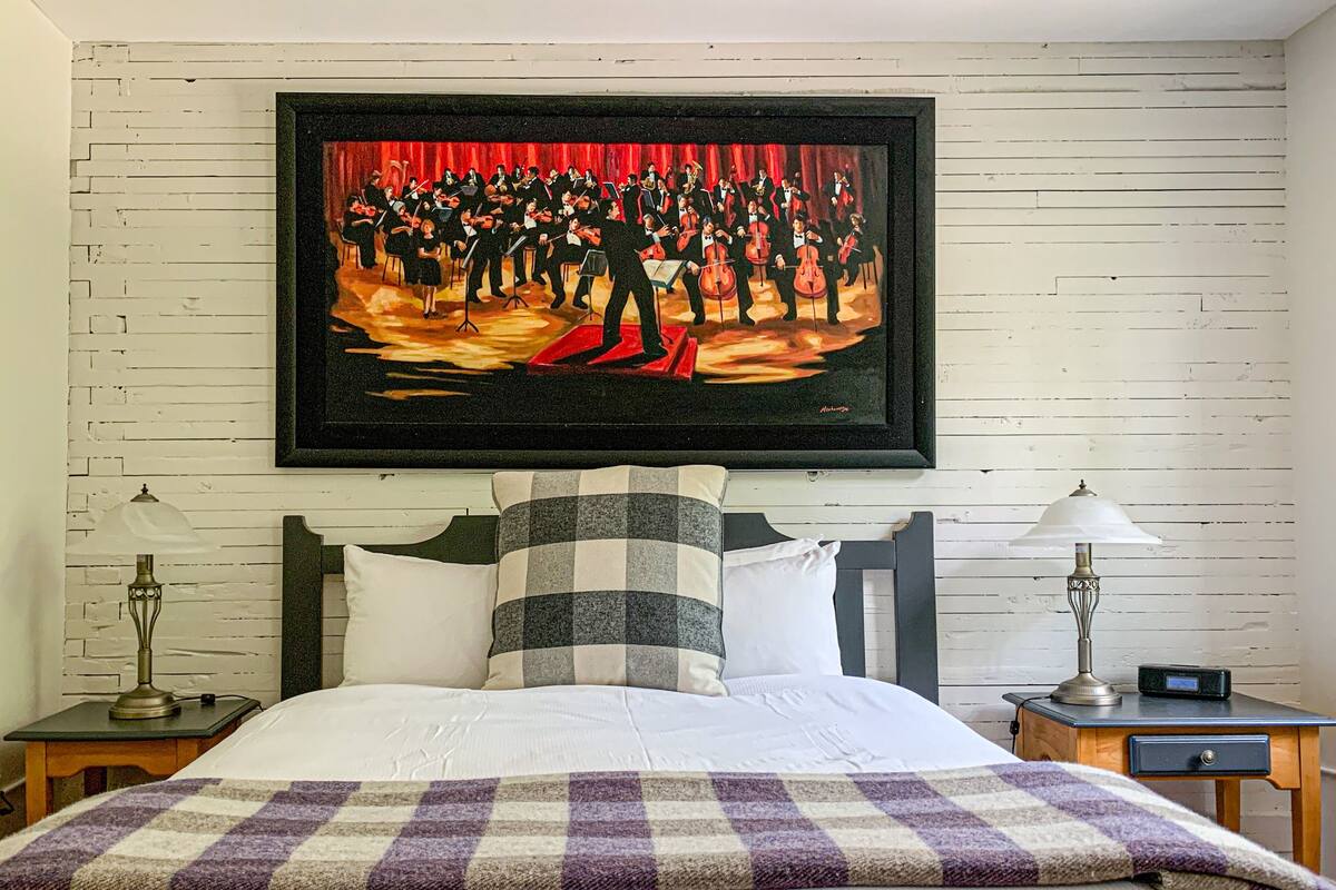 Una habitación de hotel con una cama, una manta de cuadros, una mesita de noche con una lámpara, un reloj digital y un cuadro con una banda en la pared.