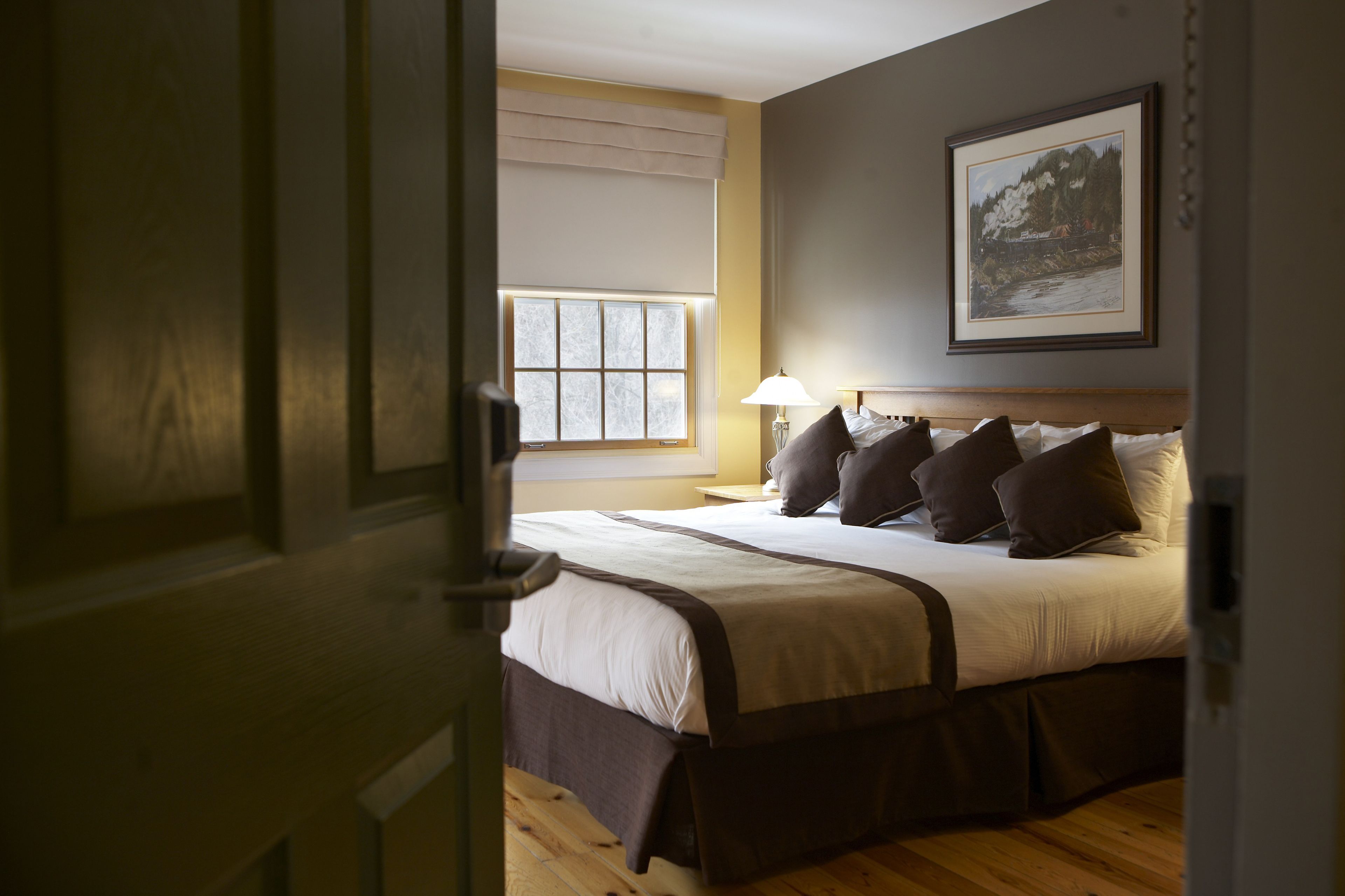 A bedroom with a bed, pillows, a nightstand, a lamp, a window, and a framed picture on the wall.
