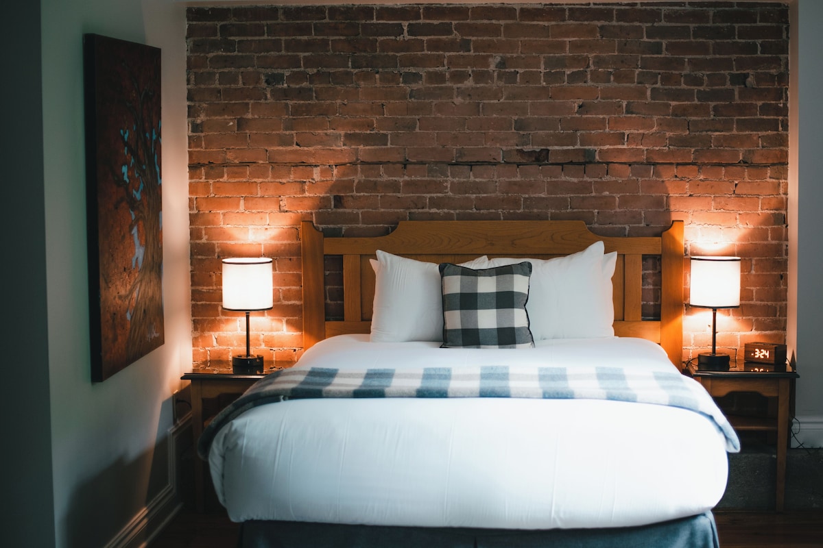 Una cama con cabecera de madera, dos lámparas de noche, un reloj digital y una pared de ladrillo al fondo.