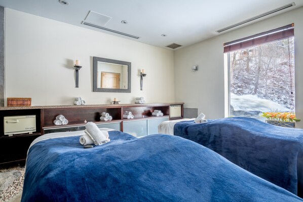 A hotel room with two beds covered in blue linens, a wooden dresser with towels, a mirror, and a window with a view of a snowy forest.