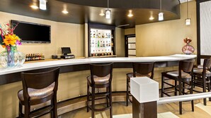 Uma área de bar com balcão curvo, vários bancos altos, uma televisão na parede e diversas garrafas expostas ao fundo.