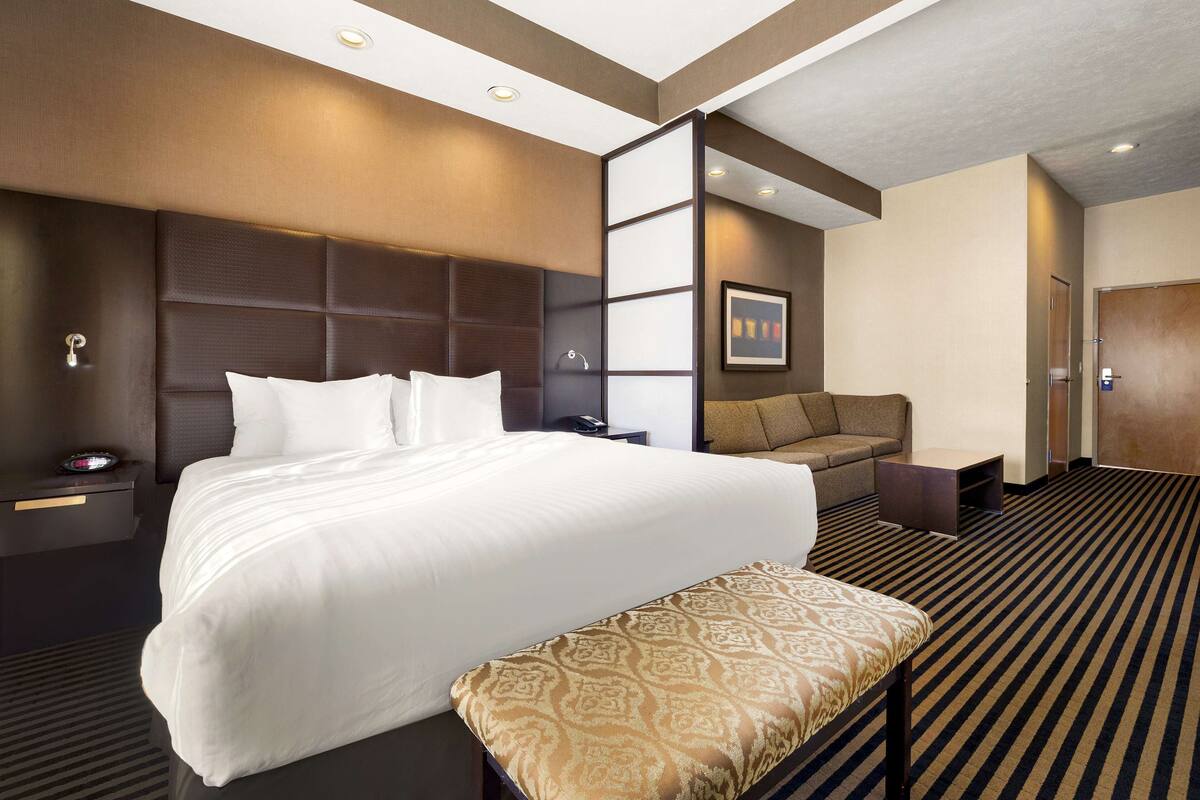 A hotel room with a large bed, a sofa, a bench, a nightstand, and a wall-mounted TV.