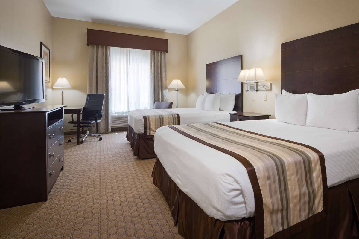 A hotel room with two beds, a desk, a chair, a TV, and a lamp.