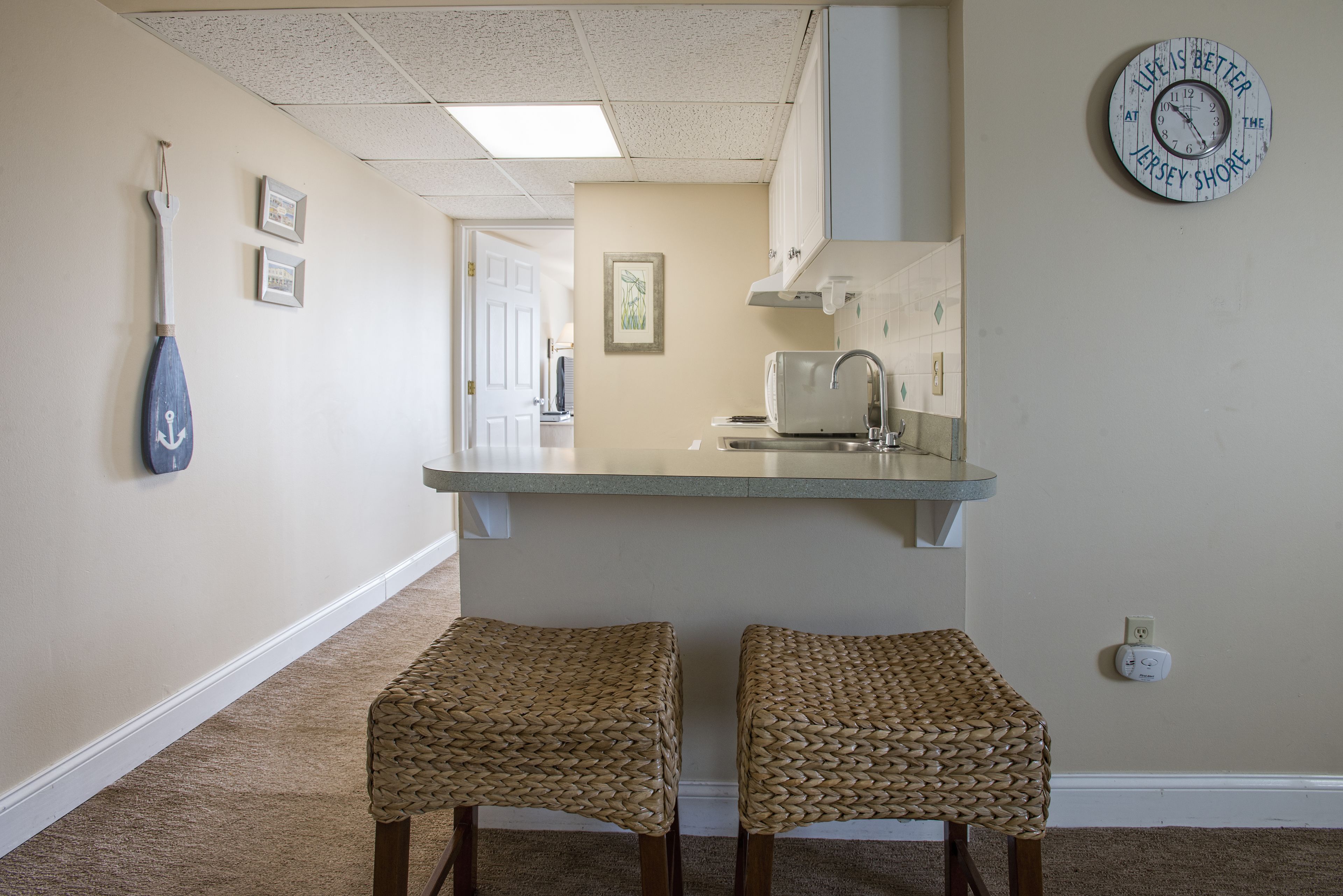 A hallway with a kitchen area, two woven stools, a clock, and framed pictures on the wall.