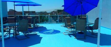 A rooftop terrace with tables and chairs, blue umbrellas, and a clear sky.