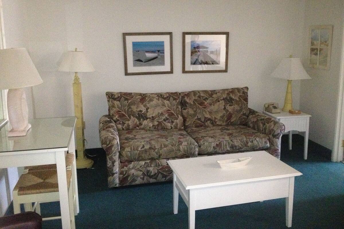 A living room with a floral sofa, a white coffee table, and two lamps. There are framed pictures on the wall.