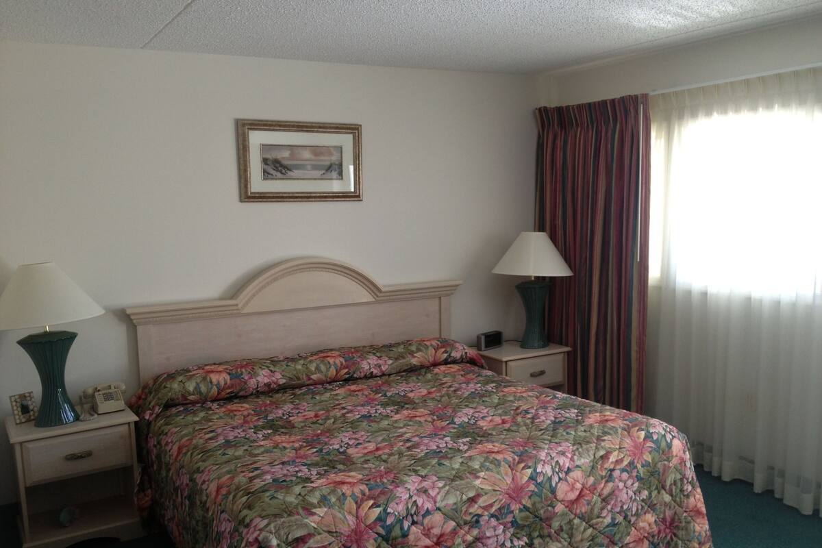 A hotel room with a floral bedspread, two bedside lamps, a framed picture on the wall, and a window with curtains.