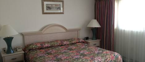 Habitación de hotel con colcha estampada con motivos florales, dos lámparas de noche, un cuadro en la pared y una ventana con cortinas.