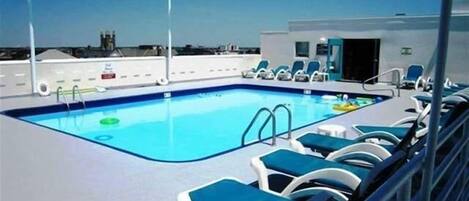 A rooftop pool with blue water, surrounded by blue lounge chairs and a few umbrellas.