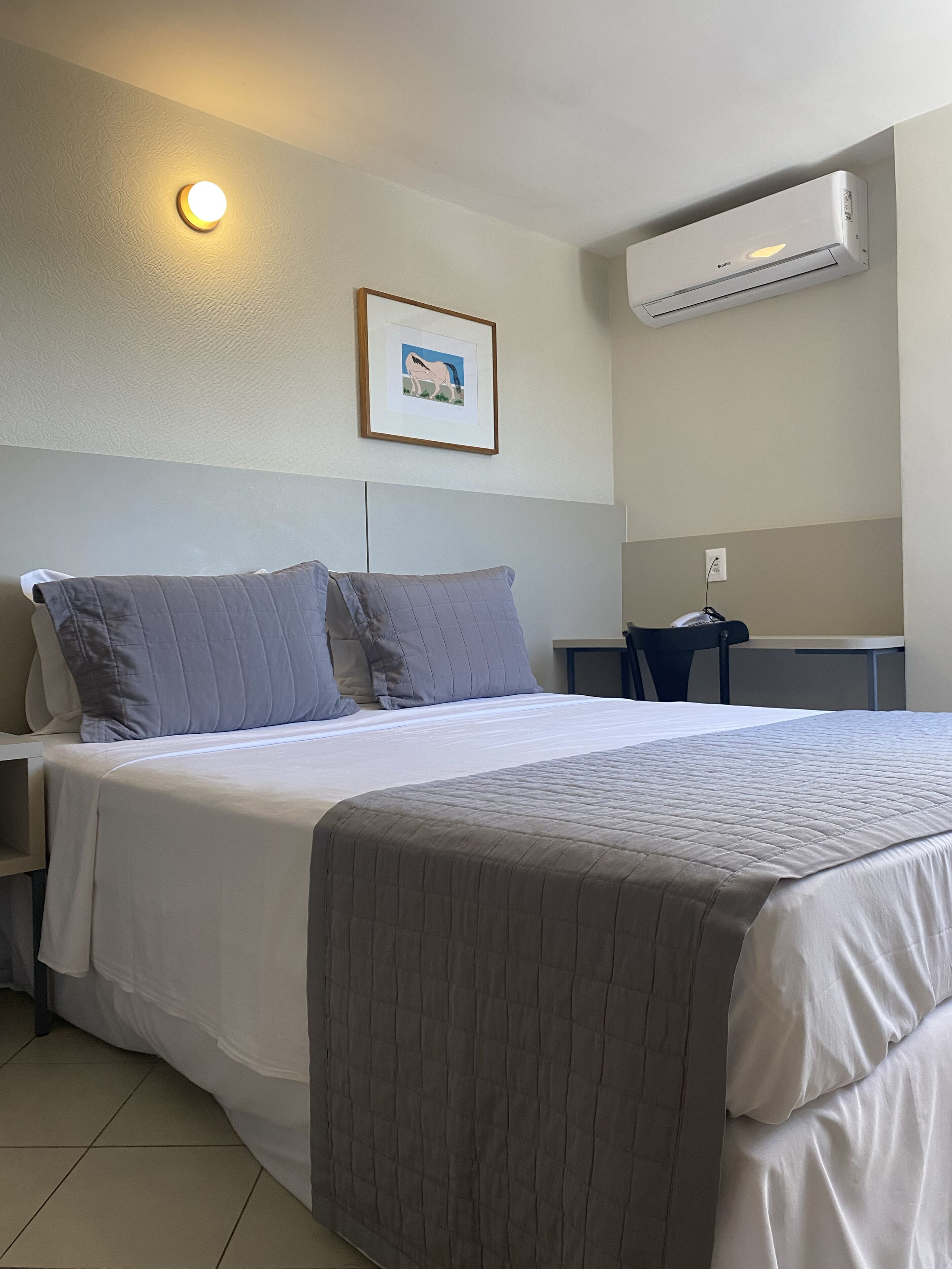 Quarto de hotel com cama, escrivaninha, ar condicionado e quadro pendurado na parede.