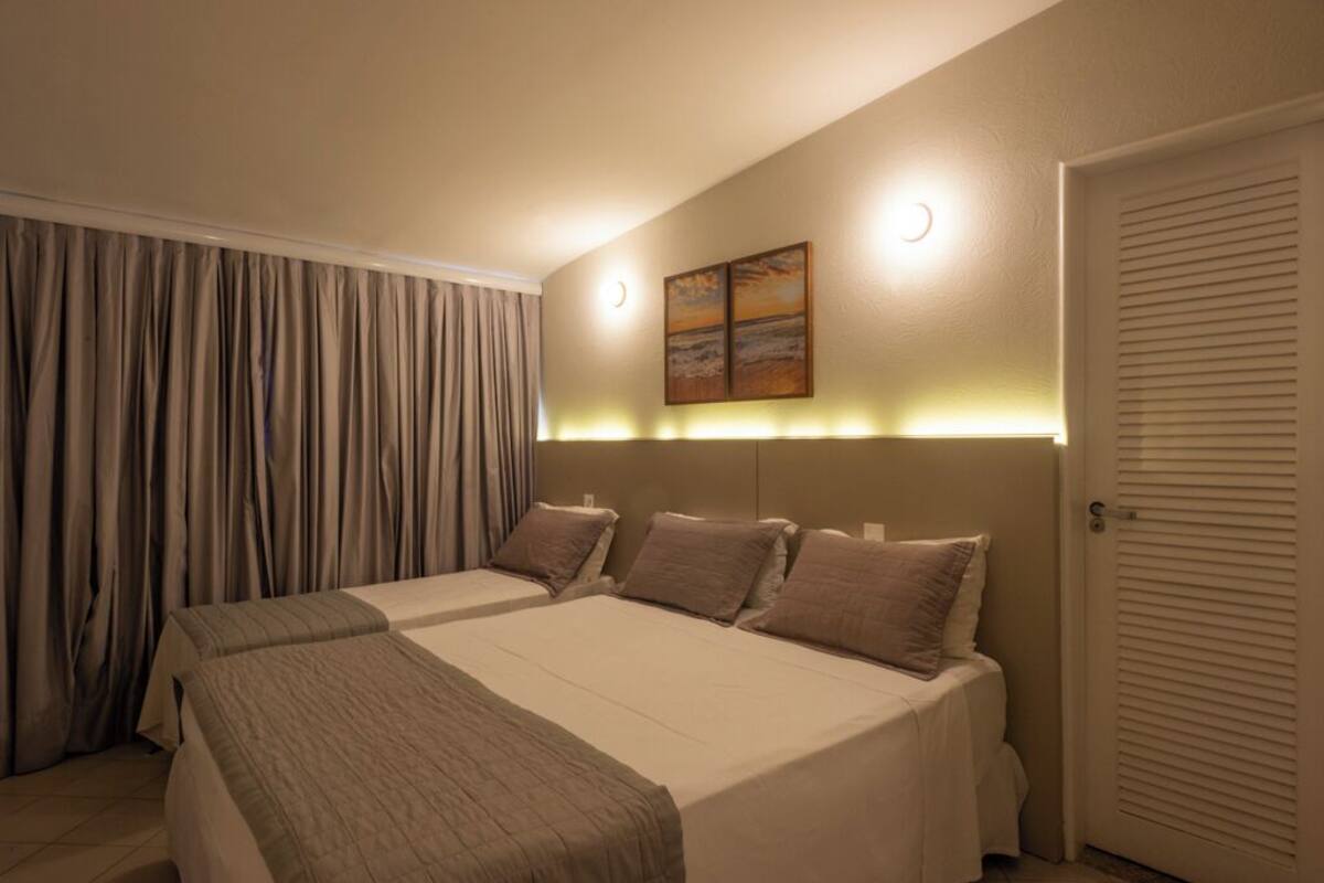 A hotel room with a large bed, a nightstand, a wall-mounted light, and a door with louvered shutters.