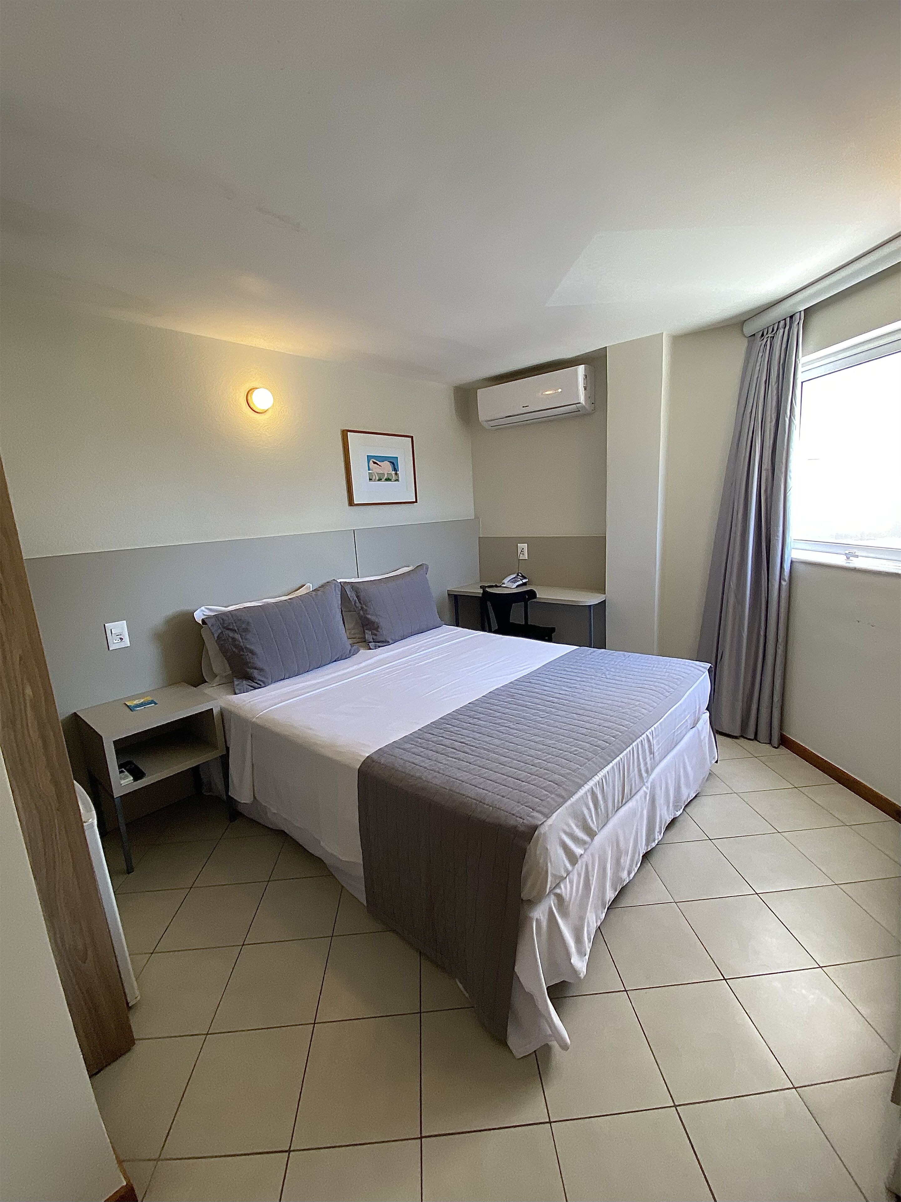 Quarto de hotel com cama, mesa de cabeceira, ar condicionado e janela com cortinas.